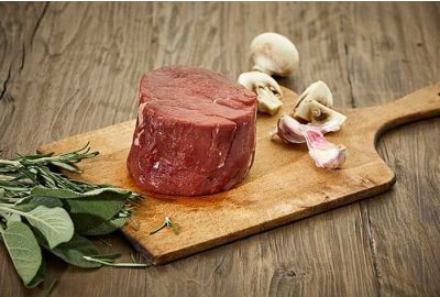 Rohes Rinderfiletsteak liegt auf einem Holzbrett, daneben frische Kräuter, Knoblauchzehen und geviertelte Champignons – alles auf einem Holztisch arrangiert.