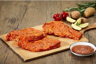 Drei gewürzte Grillhalssteaks liegen auf einem Holzbrett, bereit zum Grillen. Daneben steht eine Schale mit Marinade. Im Hintergrund sind Kartoffeln, Apfelstücke, rote Paprika und Rosmarinzweige auf einem rustikalen Holztisch zu sehen.