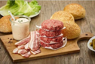 Frische Rinder-Burger Patties mit Bacon, Burgersauce, Salat und Sesam-Burgerbrötchen auf einem Holzbrett – perfekte Zutaten für selbstgemachte Burger.
