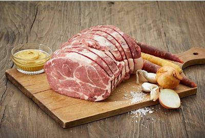 Rohes Schweinerollbratenstück vom Hals auf einem Holzbrett mit Karotten, Zwiebeln, Knoblauch und Senf – frisch aus der Metzgerei Fritsch.