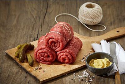 Rohe Rinderrouladen auf einem Holzbrett, daneben Gewürzgurken, Senf, Gewürze und Küchengarn zur Vorbereitung der Rouladen.