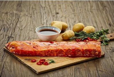 Rohes, längliches Stück marinierte Spareribs liegt auf einem Holzbrett, daneben eine Schale mit dunkler Barbecuesauce, kleine Kartoffeln, frische Kräuter und rote Chilischoten auf einem rustikalen Holztisch.