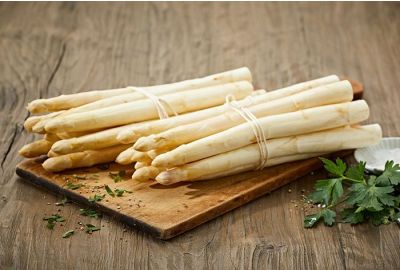 Frischer weißer Spargel liegt gebündelt auf einem Holzbrett. Daneben liegen Petersilie und eine kleine Schale mit Salz auf einem Holztisch. Metzgerei Fritsch